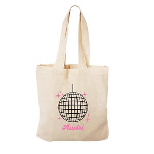 May include: A beige tote bag featuring a black disco ball graphic with pink glitter accents. The text "Audri" is printed in pink below the disco ball.
