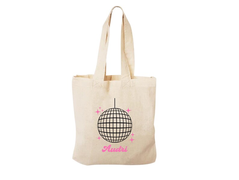 May include: A beige tote bag featuring a black disco ball graphic with pink glitter accents. The text "Audri" is printed in pink below the disco ball.