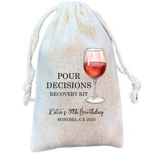 May include: A beige drawstring bag with the text "POUR DECISIONS RECOVERY KIT" and a watercolor illustration of a wine glass filled with red wine. The bag also includes the text "Katie's 30th Birthday, Sonoma, CA 2025."