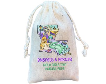 May include: A beige drawstring bag with a colorful illustration of the Louisiana state outline, featuring a green alligator, purple fleur-de-lis, and musical instruments. Text reads "Beignets & Besties, NOLA Girls Trip, August 2025."