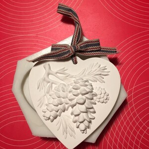 May include: White ceramic heart-shaped ornament with a raised design of pine cones and branches. The ornament has a ribbon attached for hanging.