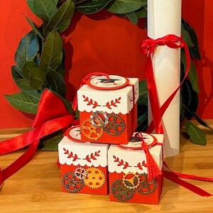 Peut inclure: Trois boîtes cadeaux rouges et blanches avec un pompon blanc et un petit motif d'engrenage. Les boîtes sont empilées les unes sur les autres. Les boîtes ont un fond blanc avec une bordure rouge et un pompon blanc. Les boîtes sont décorées d'un petit motif d'engrenage.