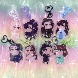 Op de afbeelding: Acht acryl sleutelhangers met chibi-stijl figuren met diverse haarkleuren en outfits, elk bevestigd aan een hartvormige sluiting in wit, paars, zwart, blauw, goud of groen, gerangschikt in twee rijen op een pastelkleurige ondergrond.
