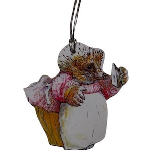 May include: A wooden Christmas bauble featuring a cartoon hedgehog wearing a pink dress and carrying a small iron. The bauble has a silver string for hanging.