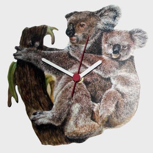 May include: A wooden wall clock depicting a koala bear and its baby on a tree branch. The clock hands are white with a red centre. The koalas are brown and grey with detailed fur.