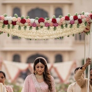 Pode incluir: Uma noiva com um traje rosa bordado caminha sob um dossel floral. O dossel é decorado com rosas vermelhas, rosas e brancas. A noiva usa joias de ouro e um adorno de cabeça. O fundo apresenta um edifício com arcos.