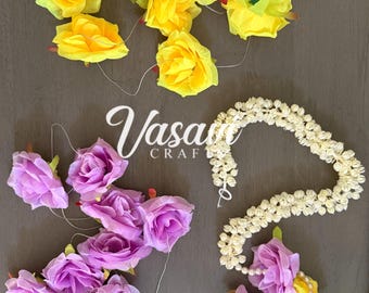 May include: A collection of artificial floral accessories including a cluster of bright yellow roses, a cluster of light purple roses, and a long garland of small white jasmine-like flowers accented with purple and yellow roses and a gold-toned pendant.