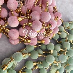 May include: Two strands of decorative trim with pink and green velvet pom-poms. Each pom-pom is attached to a gold chain with small gold beads. The trim is perfect for adding a touch of elegance to any project.