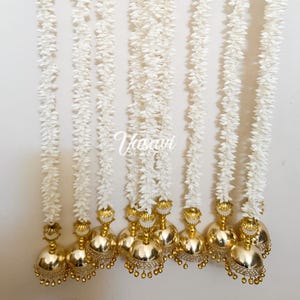 May include: Several strands of white floral garlands with gold-colored bell-shaped ornaments. The garlands are hanging against a white background. The ornaments have a decorative design with small, clear stones.