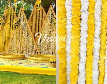 May include: A wedding ceremony backdrop decorated with yellow and white flower garlands. The garlands are strung on a wooden frame in the shape of triangles. The backdrop is set up in an outdoor setting with green grass in the foreground.