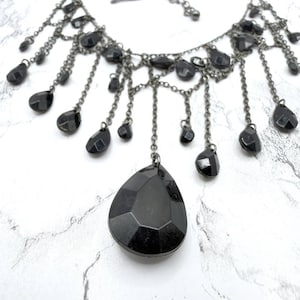 May include: A black statement necklace with a cascade of teardrop-shaped pendants. The necklace features a large, faceted teardrop pendant at the center, with smaller teardrops suspended from delicate chains. The metal has a dark, gunmetal finish.