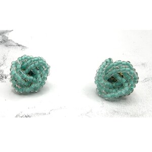 May include: A pair of turquoise beaded stud earrings. Each earring is a small, round cluster of turquoise beads, creating a textured, knotted appearance. The earrings are set against a white background.