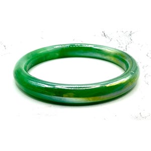 May include: A vibrant green bangle bracelet with a glossy finish. The circular bracelet has a smooth surface and a slight iridescent sheen, reflecting hints of yellow and blue. This piece of jewelry is a simple yet elegant accessory.