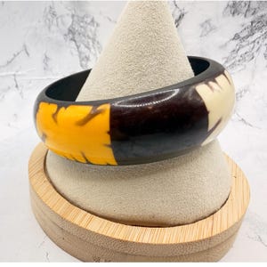May include: A thick, circular bangle bracelet featuring a polished dark brown base with contrasting sections of bright yellow and off-white cream patterns. The bracelet has a smooth, glossy finish and a wide, rounded profile that encircles the wrist.