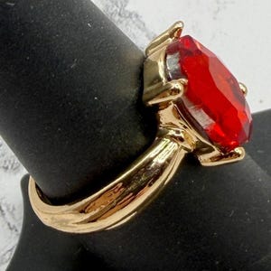 May include: A gold-colored ring featuring a large, faceted, red gemstone. The gemstone is oval-shaped and set in a gold-toned metal band. The ring is displayed on a black surface.