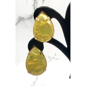 May include: A pair of teardrop-shaped earrings with a shimmering, iridescent yellow-green hue. The earrings have a gold-colored metal trim and are displayed on a black stand against a white and gray background.