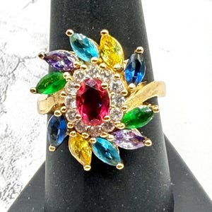 May include: A gold-toned ring featuring a central oval ruby surrounded by a halo of clear stones. The ring's design includes colorful marquise-cut gemstones in shades of blue, green, yellow, and purple, arranged in a floral pattern.