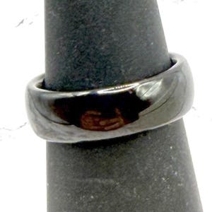 May include: A close-up of a dark gray ring with a smooth, polished surface. The ring is displayed on a black ring holder. The ring has a rounded band and a shiny, reflective finish.