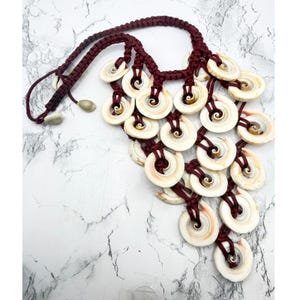 May include: A handcrafted necklace featuring a maroon braided cord and numerous white and tan shell pendants. The shell pendants are circular with a spiral design in the center, creating a unique and natural aesthetic. The necklace has a bohemian style.