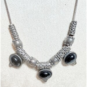 May include: A necklace featuring a silver-toned snake chain adorned with alternating cylindrical metal beads with intricate scrollwork patterns and smooth, round silver-toned beads. Three oval black cabochon stones are set as focal points.