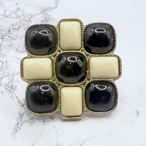 May include: A square brooch with a gold-tone metal frame. The brooch features a pattern of alternating black and cream-colored square and rectangular stones. The stones are set within the frame, creating a geometric design. The brooch is a vintage accessory.