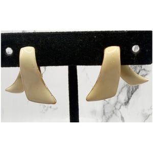 May include: A pair of cream-colored, abstract earrings. Each earring features a curved, angular design with a gold-colored trim. The earrings are displayed on a black stand against a white and gray marble background.