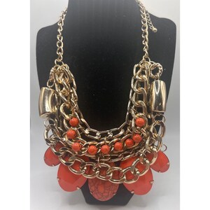 May include: A gold-toned statement necklace with multiple layers of chain and orange accents. The necklace features large, round orange pendants and smaller orange beads, creating a bold and eye-catching design.