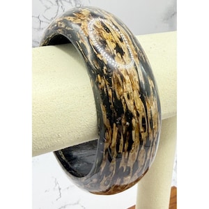 May include: A wide, polished bangle bracelet with a striking pattern of black and tan hues. The bracelet is made of a solid material and has a smooth, glossy finish. The design features a unique, organic pattern.