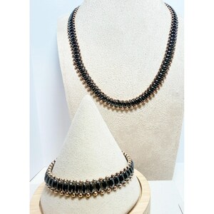 May include: A necklace and bracelet set featuring a repeating pattern of dark, metallic, oval-shaped beads interspersed with small, round, gold-toned metallic beads. The jewelry pieces are constructed with a consistent, uniform design throughout.