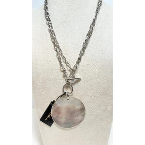 May include: A silver-toned necklace featuring a long chain with a toggle clasp and a large, circular, iridescent shell pendant. The pendant displays natural grey and white swirling patterns, and a small black rectangular tag is attached to the ring.