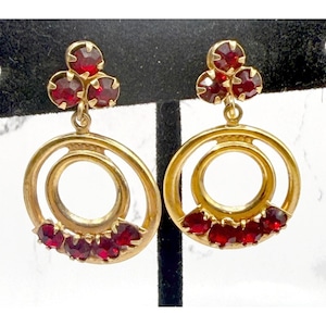 May include: Gold-toned earrings with a cluster of three red gemstones at the top and a double-circle design. The lower portion of the earrings features a row of red gemstones. The earrings are displayed on a black stand.