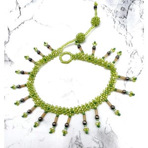 May include: A beaded necklace in shades of green, with a delicate design. The necklace features small, round beads and elongated accents, creating a unique, eye-catching accessory. The necklace is displayed on a white surface.