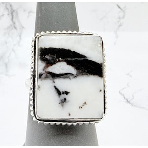 May include: A rectangular ring featuring a white stone with prominent black inclusions. The stone is set in a silver-toned metal bezel with a decorative notched edge, creating a distinct contrast between the light and dark patterns on the surface.