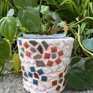 May include: A cylindrical plant pot with a textured light pink base decorated with a mosaic pattern of small, multi-colored geometric tiles in shades of white, blue, grey, brown, and terracotta, containing a green leafy plant with heart-shaped leaves.