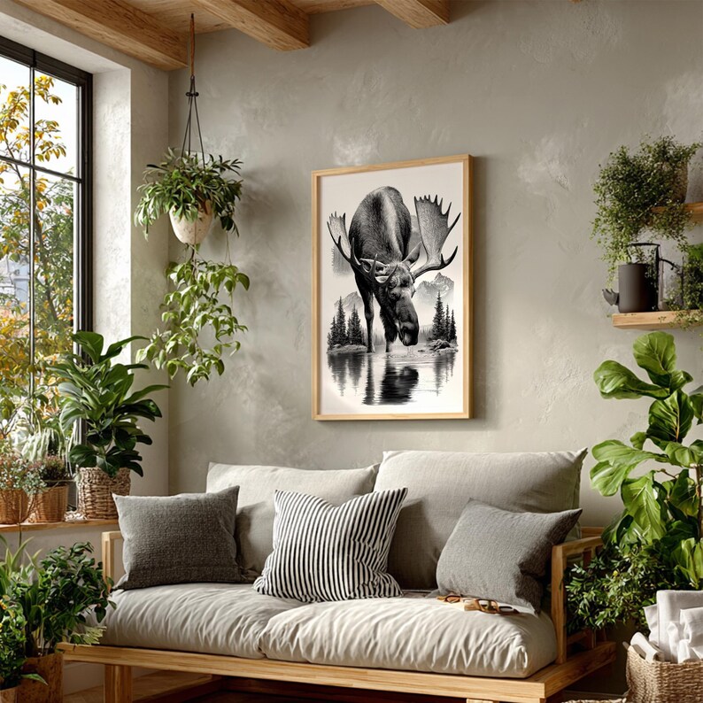 May include: A framed black and white print of a moose drinking from a lake, set against a backdrop of mountains and trees. The artwork is displayed above a light-colored sofa with decorative pillows, creating a cozy living room scene.