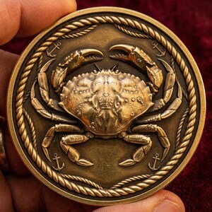 Op de afbeelding: Een bronzen munt met een gedetailleerd krabontwerp in het midden. De krab is omgeven door een touwachtige rand met ankers. De munt heeft een verweerde, antieke uitstraling.