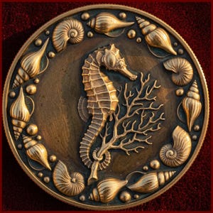 May include: A circular bronze-toned medallion featuring a central relief of a seahorse grasping a piece of coral. The border is decorated with various embossed seashells and small spheres arranged in a circular pattern around the center.