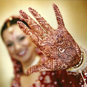 以下が含まれることがあります： インドの伝統的な結婚式で用いられる、複雑なヘナ模様が施された女性の腕。ヘナは濃い赤褐色で、指先から手のひらまで全体を覆っています。女性は赤と金の衣装を着ており、背景では顔がぼやけています。