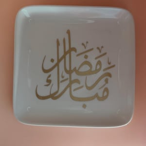 May include: A square, white ceramic tray with rounded corners. The tray features gold Arabic calligraphy in the center. The background is a solid, light peach color.