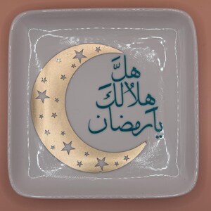May include: A square, off-white ceramic dish with a gold crescent moon and star cutouts. Teal Arabic calligraphy is centered on the dish. The dish has rounded corners and a glossy finish.