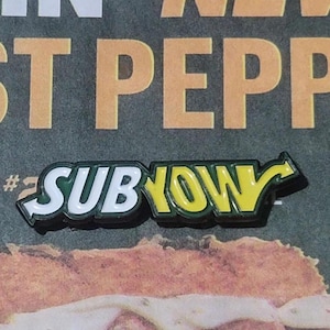 May include: A green and yellow enamel pin with the word "SUBWAY" in white letters and a yellow arrow pointing to the right.