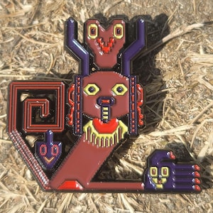 以下が含まれることがあります： 茶色の体、紫色の角、黄色と赤のアクセントが特徴の、様式化された人物を描いた詳細なピン。 デザインには幾何学的な形と小さな様式化された手が含まれています。 ピンは乾燥した草の背景に設定されています。