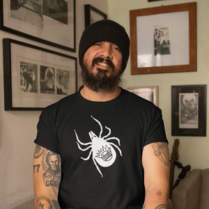 May include: A man wearing a black beanie and a black t-shirt with a white graphic of a spider with a crown on its head. He is smiling and looking at the camera.