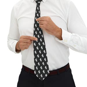 May include: A black necktie featuring a repeating white graphic pattern of a person playing an electric guitar. The tie is worn over a crisp white long-sleeved button-down shirt and is paired with dark trousers held up by a dark belt.