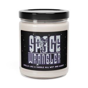 Puede incluir: Un tarro de vidrio para velas con tapa negra y una etiqueta que dice "Space Wrangler" con un gráfico de astronauta. La etiqueta también dice "Huele como una silla de montar toda mojada y caliente."