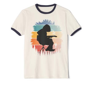 May include: Cream-coloured ringer t-shirt with a navy blue collar and sleeve trim. The shirt features a black silhouette of a person playing a guitar, set against a colourful horizontal striped background.