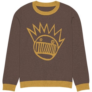 May include: A brown long-sleeved sweater with a yellow ribbed crew neck and cuffs. The sweater features a yellow graphic of a cartoon face with a crown of spikes.