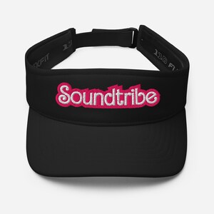 May include: Black visor with the embroidered text "Soundtribe" in white with a pink outline. The visor has a curved brim and an adjustable strap. The text is in a stylized font.