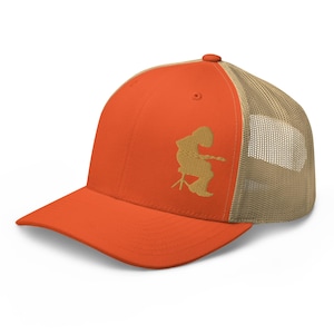 May include: Orange trucker cap with a tan mesh back and a curved peak. The front of the cap features a gold embroidered design of a person playing a stringed instrument. The cap has a snapback closure.