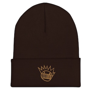 May include: A brown knitted beanie with a gold embroidered logo of a cartoon face with a crown on top.